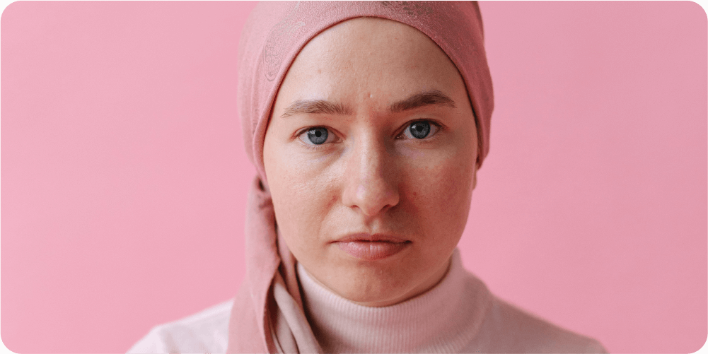 Человек в розовом платке и свитере пристально смотрит в камеру. Фон однотонно-розовый, создающий спокойную и сосредоточенную атмосферу.