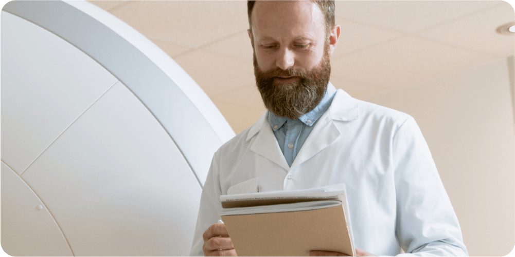 Врач с бородой в белом халате просматривает документы в медицинском учреждении. На заднем плане — аппарат МРТ, что говорит о профессионализме.
