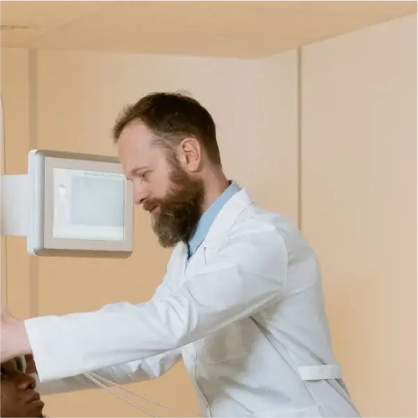 Врач в белом халате настраивает медицинское оборудование рядом с пациентом в клинике. Нейтральные тона помещения создают спокойную и профессиональную атмосферу.