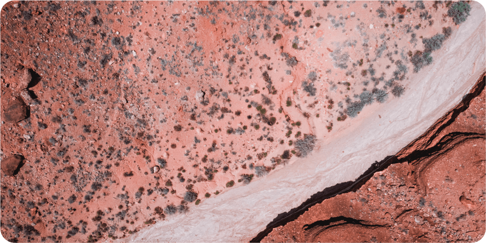 Вид с воздуха на каменистый пустынный ландшафт с красно-белыми полосами. Редкая растительность усеивает местность. Сцена создаёт ощущение засушливого одиночества.