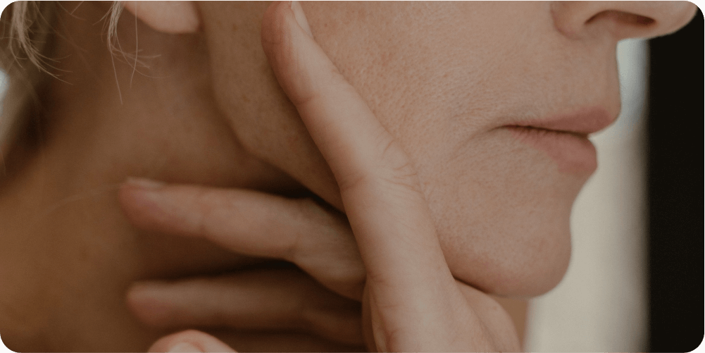 Крупный план подбородка и губ человека, рука которого нежно касается его шеи и линии подбородка, передавая самоанализ и спокойствие посредством мягкого освещения и естественной текстуры кожи.